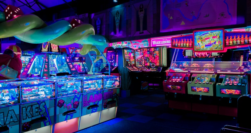 An indoor arcade filled with vibrant gaming machines lit with neon blue and pink lights. The scene includes various arcade games with flashing screens and colorful displays, and a large overhead structure resembling a green snake. The background shows illuminated signs and themed decor, contributing to an immersive entertainment atmosphere.