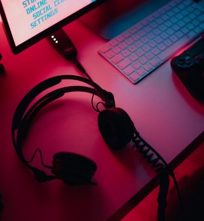 A dimly lit gaming setup with a monitor displaying a game's menu on the screen. A mechanical keyboard sits in front of the monitor. A pair of over-ear headphones and a game controller rest on the desk. The scene is bathed in a red and blue light, creating a moody gaming atmosphere.