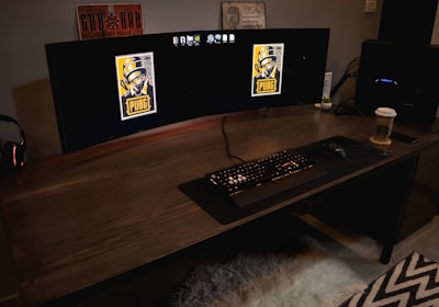 A gaming setup featuring a large curved monitor displaying PUBG logos on the screens. A keyboard and mouse are positioned on a large desk pad. A pair of headphones rests on the desk alongside a mobile phone. A drink in a Starbucks cup is placed to the right, while a desktop tower is seen in the background on the right side.