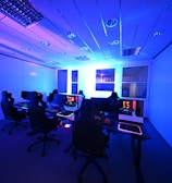 A modern gaming setup with multiple high-backed gaming chairs and monitors arranged in a room with a cool blue lighting. The computers have illuminated keyboards and glowing screens, creating a high-tech ambiance.