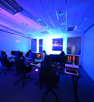 A modern gaming setup with multiple high-backed gaming chairs and monitors arranged in a room with a cool blue lighting. The computers have illuminated keyboards and glowing screens, creating a high-tech ambiance.