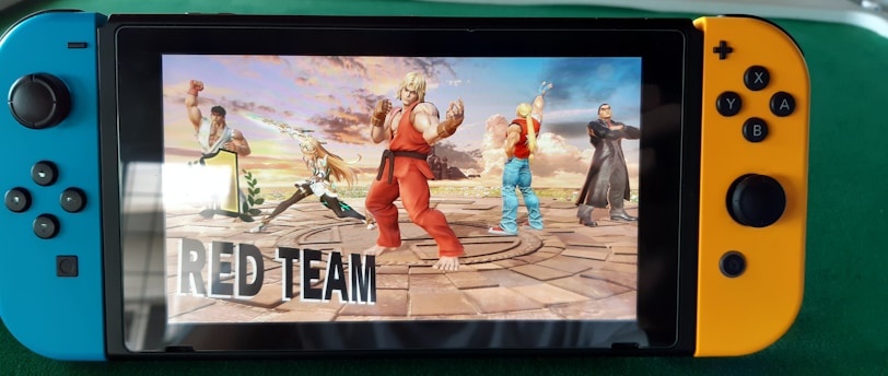 A handheld gaming console displays a screenshot of a fighting game featuring cartoonish characters in dynamic poses. The characters are standing in a virtual arena setting with a sky and cloud backdrop. The console is placed on a green surface.