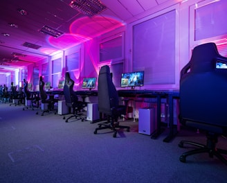 A row of gaming computers with large monitors and racing-style chairs is set up along the interior of a modern room. The space is illuminated with vibrant purple and blue LED lighting, creating a futuristic atmosphere. The room is organized and clean, suggesting a professional gaming or esports environment.
