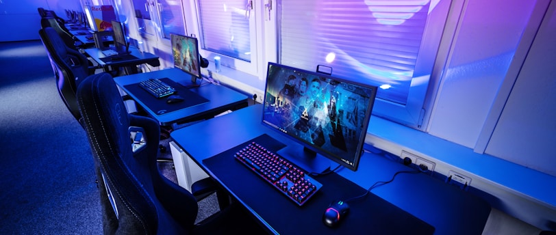A row of modern gaming setups is arranged neatly along a window-lined wall. Each station includes a high-resolution monitor displaying vivid graphics, a mechanical keyboard with colored lighting, a gaming mouse, and an ergonomic chair. The room is illuminated with ambient blue and purple lights, creating a sleek and futuristic atmosphere.