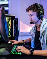 A young person is intensely focused on a computer screen while wearing a headset with a microphone. They are using a gaming keyboard and mouse, sitting in a gaming chair. The room has cool lighting, with a visible PC tower.