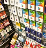 A display rack featuring a variety of gift and prepaid cards from popular brands. Cards are organized in stacks and rows, including Google Play, Apple iTunes, Amazon, PlayStation, Netflix, and more. The colors are vibrant and the packaging is clearly branded.