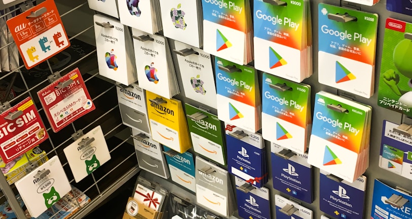 A display rack featuring a variety of gift and prepaid cards from popular brands. Cards are organized in stacks and rows, including Google Play, Apple iTunes, Amazon, PlayStation, Netflix, and more. The colors are vibrant and the packaging is clearly branded.