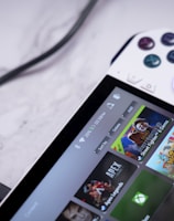 A handheld gaming console with a white body and colorful buttons features a screen displaying various game icons. The device is connected to a power source via a black cable and rests on a marble surface. The interface shows games like Apex and Street Fighter.