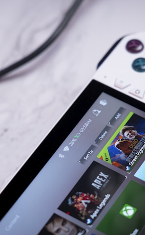 A handheld gaming console with a white body and colorful buttons features a screen displaying various game icons. The device is connected to a power source via a black cable and rests on a marble surface. The interface shows games like Apex and Street Fighter.